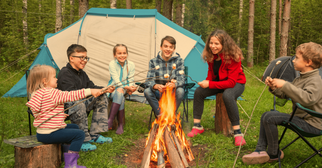 A group of children sitting around a campfire roasting marshmallows with a tent behind them